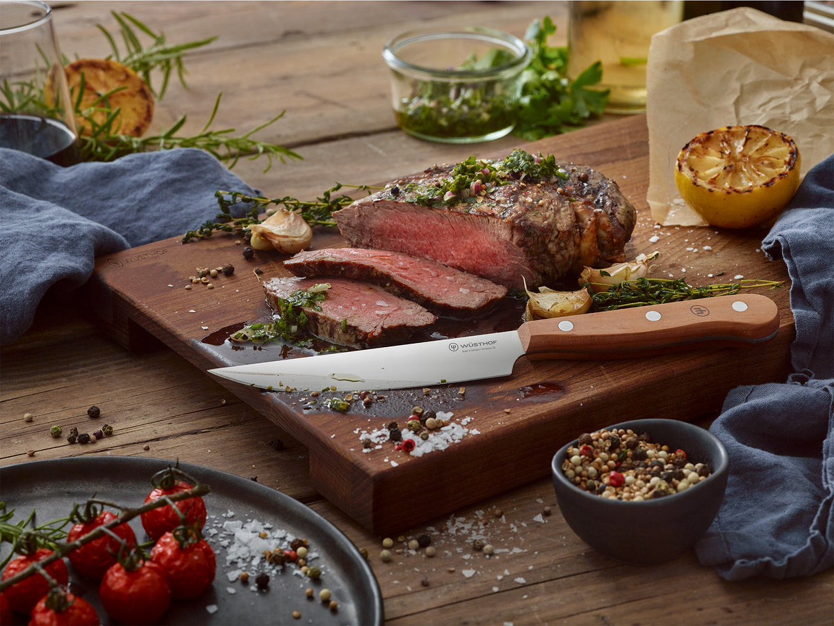 4 1/2" Plum Wood Steak Knife with a sharp stainless steel blade and smooth plum wood handle placed on a wooden board with sliced steak and herbs.