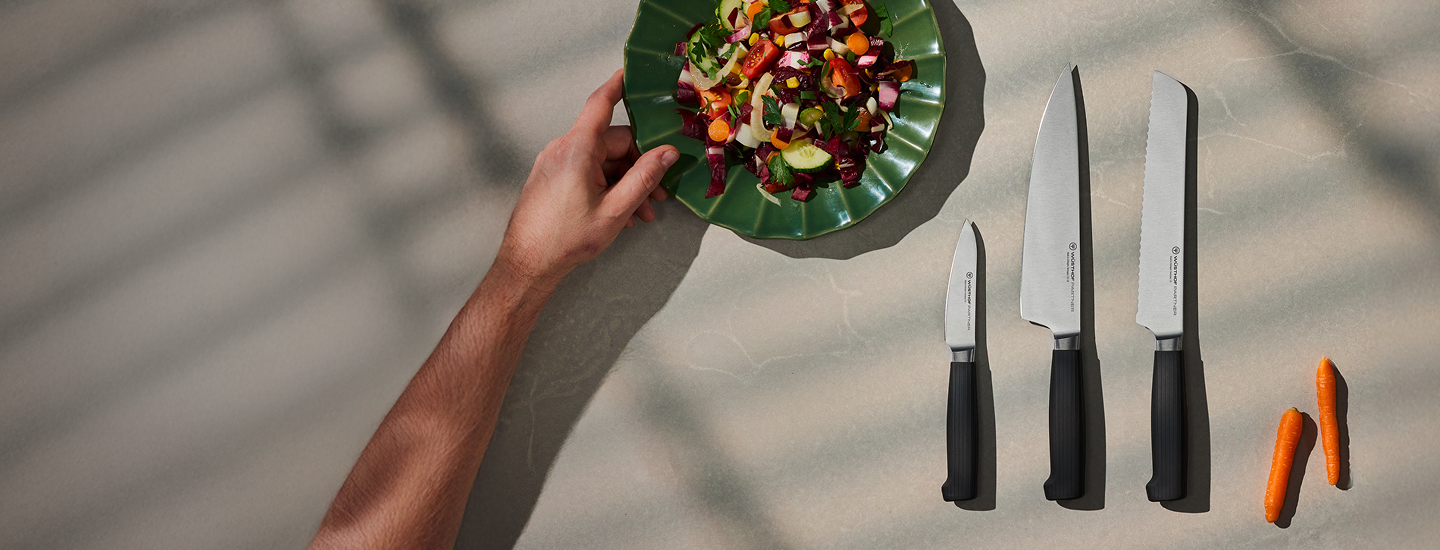 A hand holding a green plate with a colorful salad beside three black-handled kitchen knives of varying sizes and two small orange carrots on a light surface.