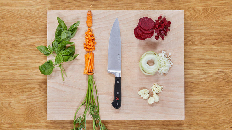 Classic 8" Chef's Knife with a full tang and black handle placed on a wooden cutting board surrounded by fresh chopped vegetables and herbs.