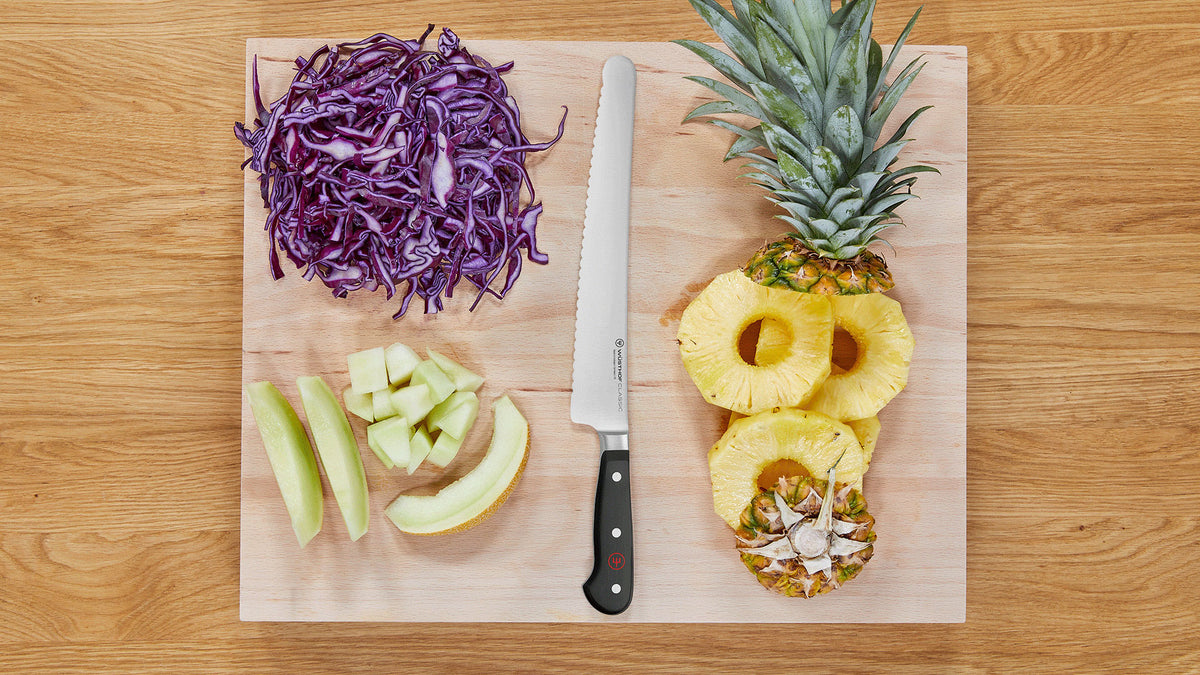 Classic 10" Super Slicer with a long serrated blade and triple-riveted handle placed on a board with sliced pineapple, cabbage, and melon wedges.