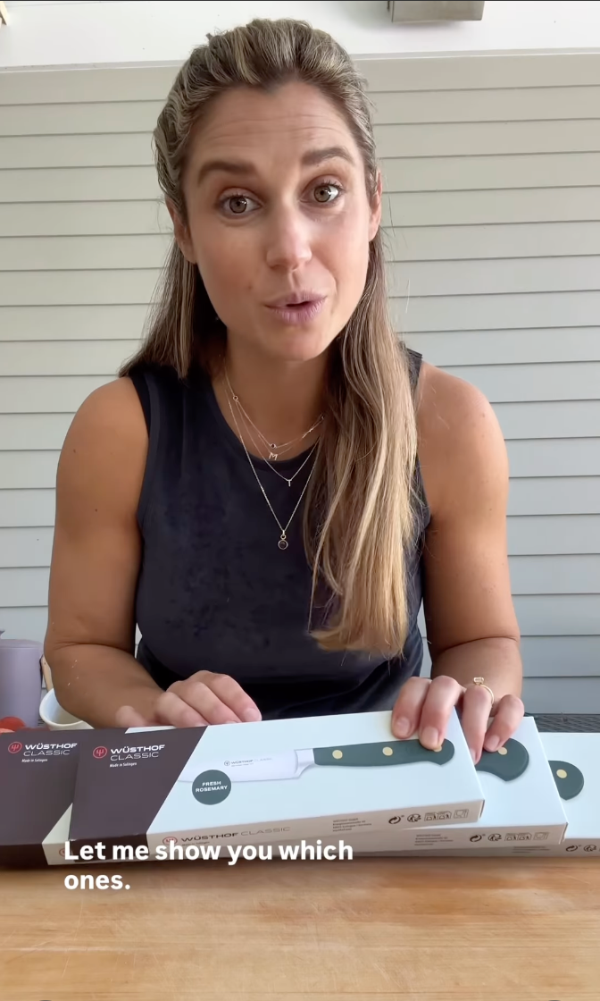 Woman with long hair wearing a sleeveless black top sitting at a wooden table with three boxed WÜSTHOF knives, speaking with text overlay reading "Let me show you which ones.