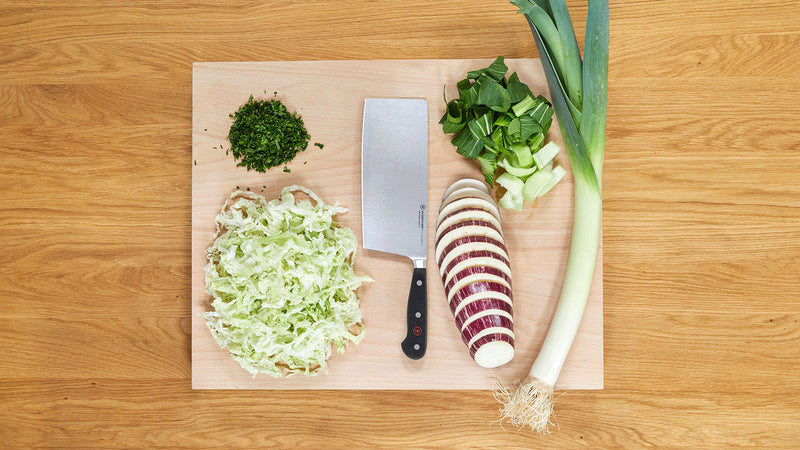 Classic 7" Chinese Chef's Knife with wide stainless steel blade placed on wooden cutting board alongside chopped green herbs, cabbage, sliced eggplant, and whole leek.