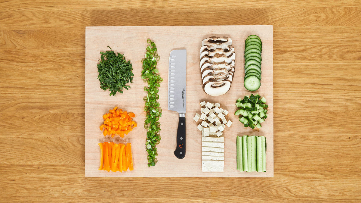 Classic 7" Hollow Edge Nakiri with a triple-riveted black handle placed on a wooden cutting board surrounded by neatly chopped vegetables including mushrooms, cucumber, tofu, and bell pepper.