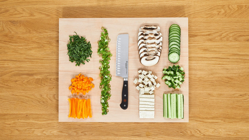 Classic 7" Hollow Edge Nakiri with a triple-riveted black handle placed on a wooden cutting board surrounded by neatly chopped vegetables including mushrooms, cucumber, tofu, and bell pepper.