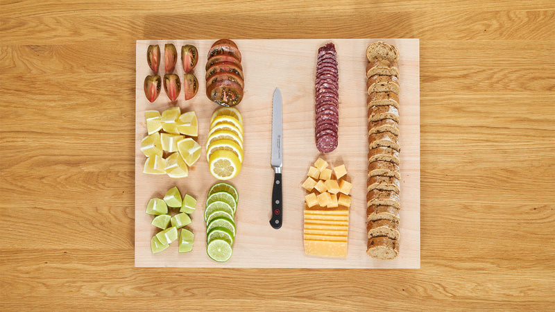 Classic 5" Serrated Utility Knife with a black riveted handle positioned on a wooden cutting board surrounded by sliced fruits, cheese, and crackers.