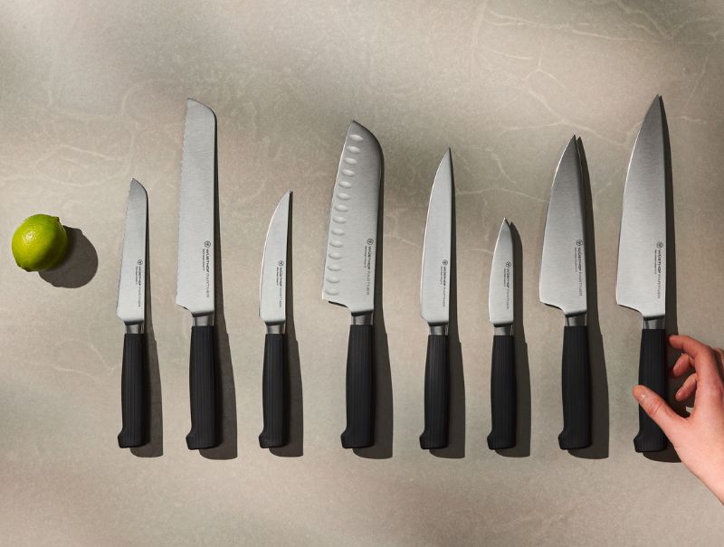 A set of nine kitchen knives with black handles arranged in a row on a light countertop, with a lime on the left and a hand holding the largest knife on the right.