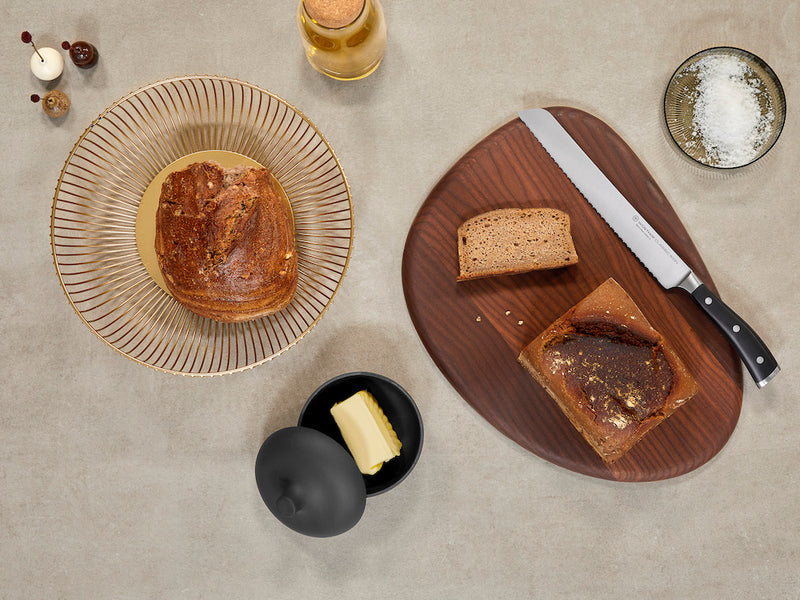 Serrated knife with black handle resting on a wooden cutting board beside sliced brown bread, a plate with a whole bread loaf, butter in a black dish with lid, a bowl of coarse salt, and a glass bottle on a beige countertop.