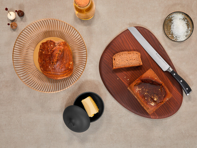 Classic Ikon 9" Precision Double-Serrated Bread Knife with a forged German steel blade and a black handle displayed next to sliced bread on a wooden board.