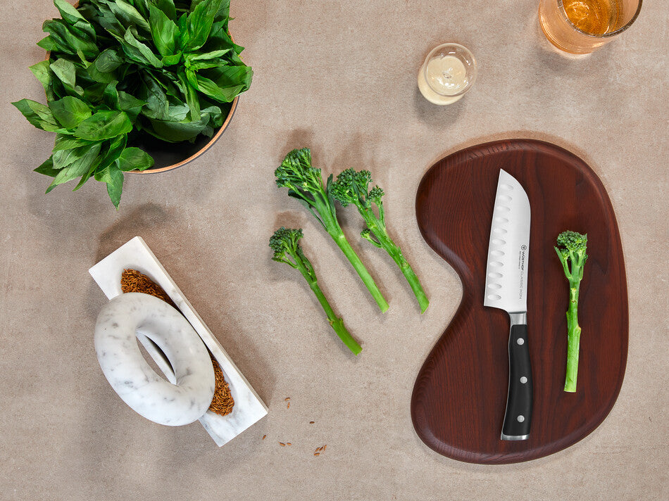 Classic Ikon 2-Piece Santoku Set featuring a forged German Santoku knife with a black ergonomic handle displayed on a wooden board alongside fresh broccoli rabe and a kitchen setting.