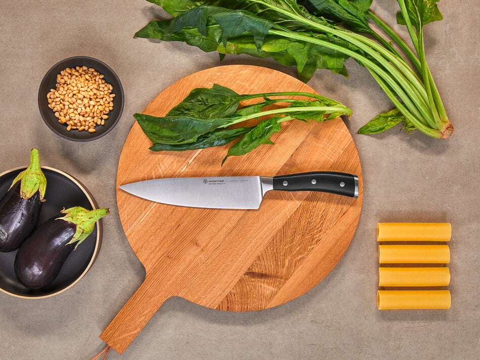 Classic Ikon 8" Chef's Knife with a forged German steel blade and triple-riveted black handle placed on a wooden paddle cutting board with fresh greens.