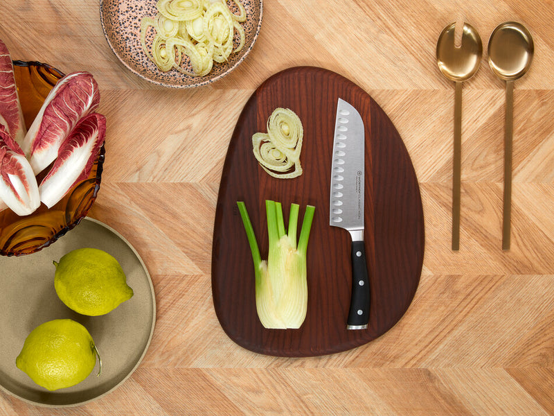 Classic Ikon 7" Hollow Edge Santoku with forged German steel blade and triple-riveted handle displayed on a wooden board with fresh fennel and sliced onions.