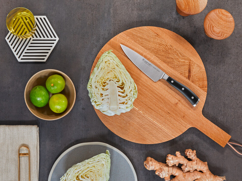 Classic Ikon 6" Chef's Knife with a forged German steel blade and ergonomic triple-riveted black handle resting on a round wooden cutting board.