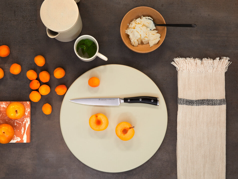 Classic Ikon 6" Utility Knife with a forged German steel blade and a triple-riveted black handle displayed with peeled apricots on a round cutting board.