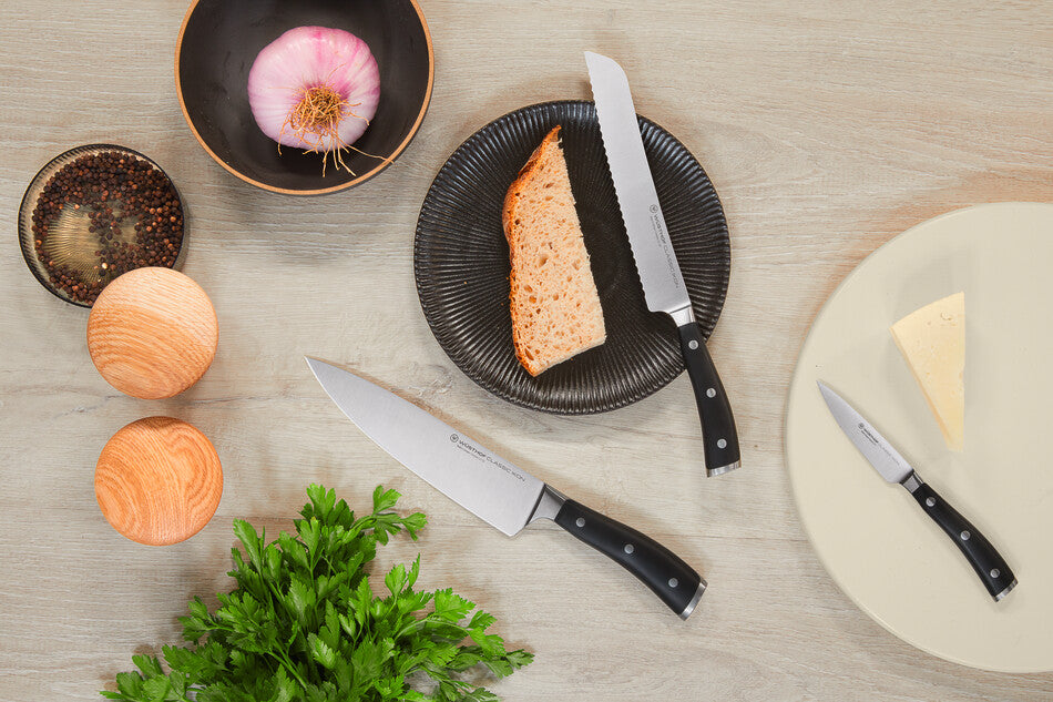 Classic Ikon 3-Piece Knife Set featuring forged German steel blades with black ergonomic handles displayed with fresh herbs, garlic, bread, and cheese for kitchen use.