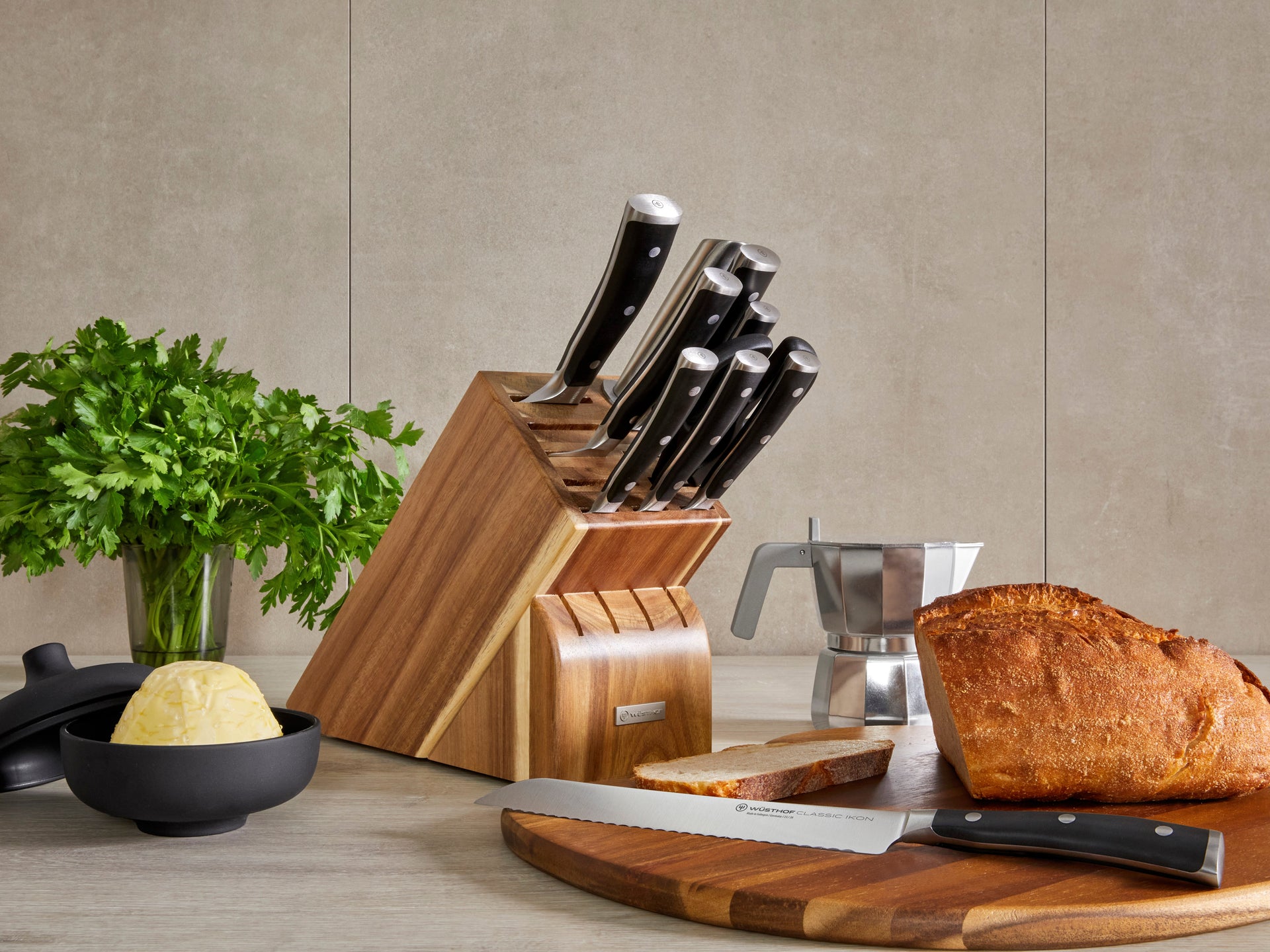 Set of knives in a wooden block on a kitchen counter with bread and herbs.