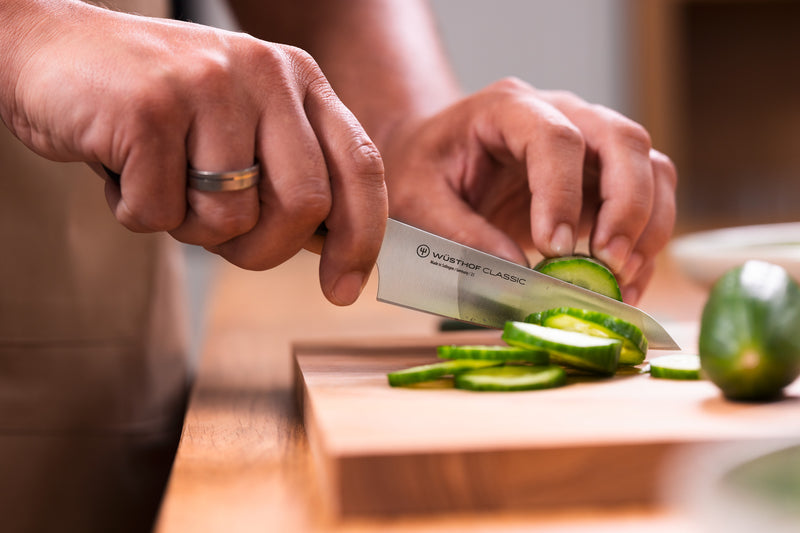 Classic 4 1/2" Asian Utility Knife with a forged German steel blade slicing fresh cucumber on a wooden cutting board in a home kitchen setting.