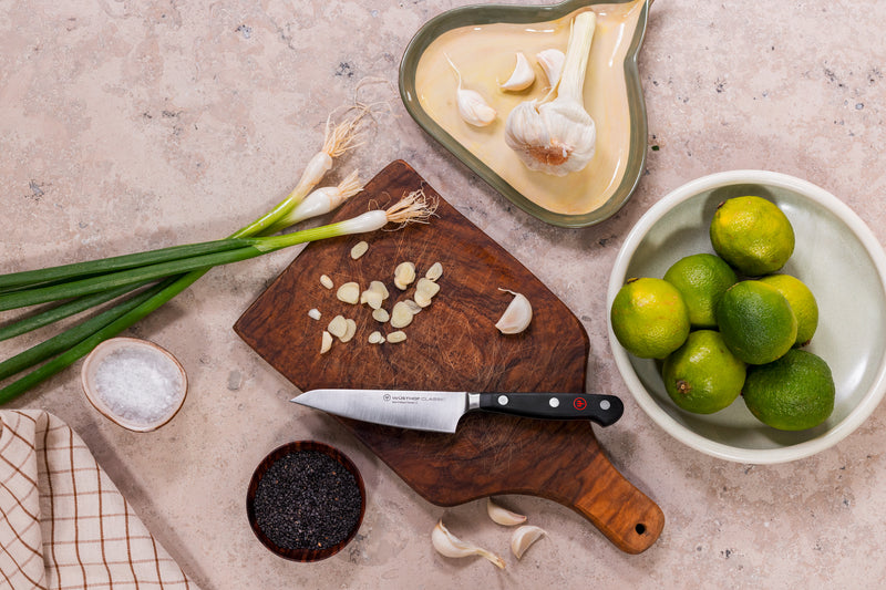 Classic 4 1/2" Asian Utility Knife with a forged German steel blade and triple-riveted handle resting on a wooden board with chopped garlic, green onions, limes, and coarse salt nearby.