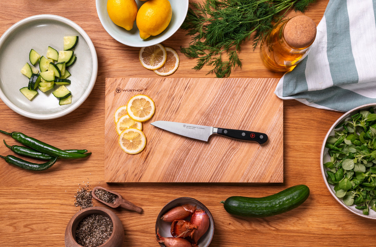 Classic 4 1/2" Asian Utility Knife with forged German steel blade and triple-riveted handle resting on a wooden board with sliced lemons surrounded by fresh ingredients.