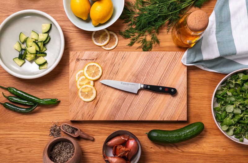 Classic 4 1/2" Asian Utility Knife with forged German steel blade and triple-riveted handle resting on a wooden board with sliced lemons surrounded by fresh ingredients.