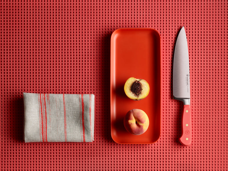 Classic 8" Chef's Knife with a polished forged German steel blade and vibrant coral handle, displayed beside halved peaches on a red tray and folded striped cloth.