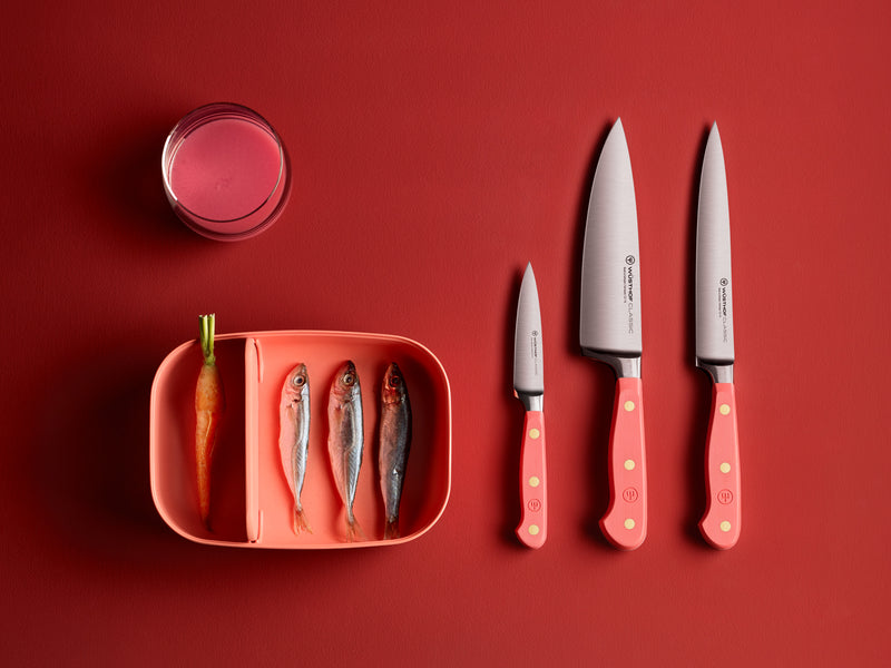Classic 3 1/2" Paring Knife with a smooth coral handle featuring three brass rivets and a polished forged German steel blade, displayed alongside matching knives and ingredients.