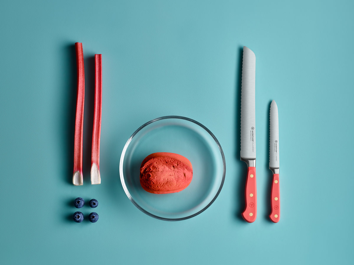 Classic 5" Serrated Utility Knife with a bright red handle and brass rivets displayed next to fresh rhubarb stalks, blueberries, and a red potato in a glass bowl on a blue background.