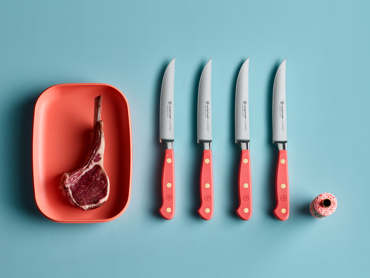 Four Classic 4 1/2" Steak Knives with coral-colored handles and brass rivets lined up next to a raw tomahawk steak in an orange dish on a blue background.