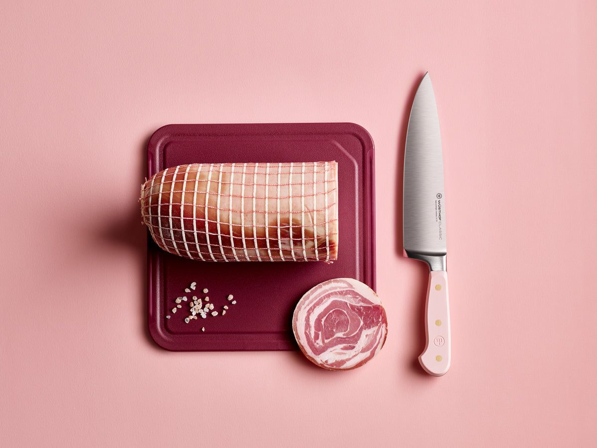 Classic 8" Chef's Knife with a polished forged German steel blade and pale pink handle resting beside a cutting board holding a netted raw roast and sliced pork belly.