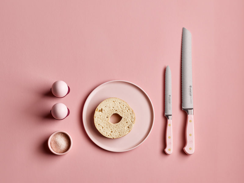 Classic 5" Serrated Utility Knife set with pale pink handles featuring subtle gold dots, displayed alongside a sliced bagel, pink salt, and matching salt and pepper shakers on a pink background.
