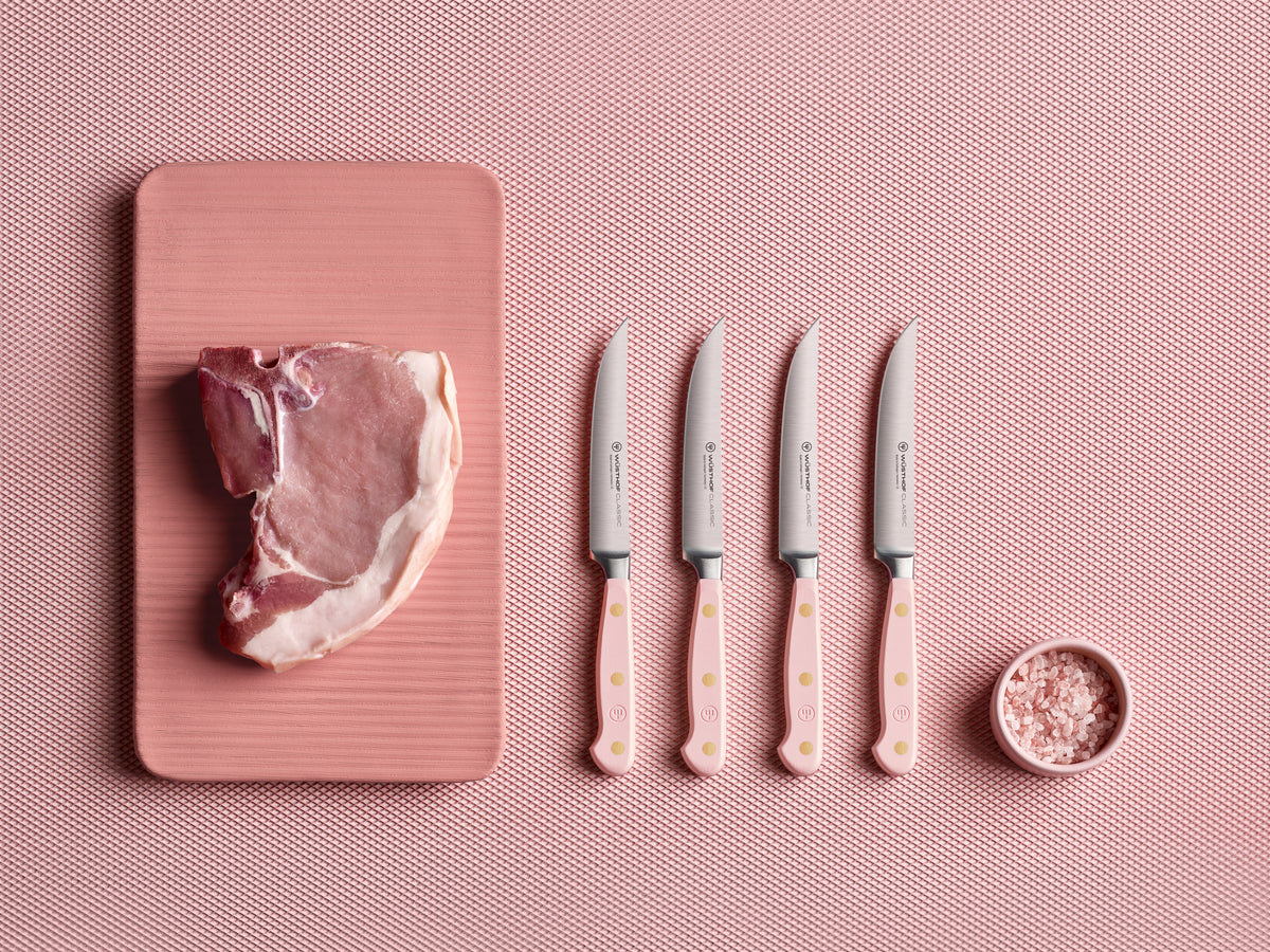 Classic 4-Piece Steak Knife Set featuring light pink ergonomic handles with three brass rivets and sharp forged German steel blades displayed next to raw pork chop and pink salt.