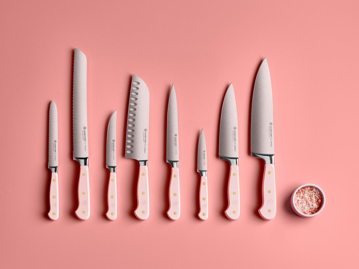 Classic 8" Chef's Knife and matching set of forged German steel kitchen knives with pale pink handles arranged in a row next to a small dish of pink salt on a pink background.