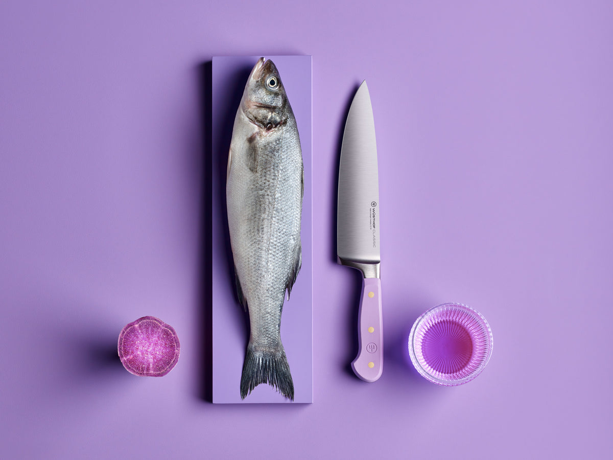 Classic 8" Chef's Knife with a polished forged German steel blade and lavender handle featuring three brass rivets, displayed beside a fresh whole fish and purple produce on a purple background.