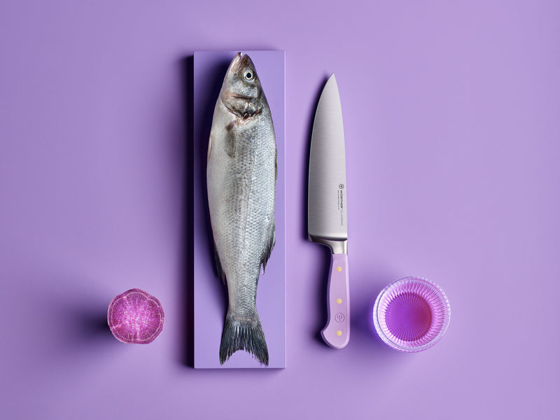 Classic 8" Chef's Knife with a polished forged German steel blade and lavender handle featuring three brass rivets, displayed beside a fresh whole fish and purple produce on a purple background.