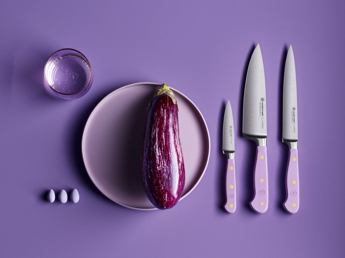Classic 6" Chef's Knife set with forged German steel blades and lavender handles featuring brass rivets, displayed alongside an eggplant, three eggs, and a glass of water on a purple background.