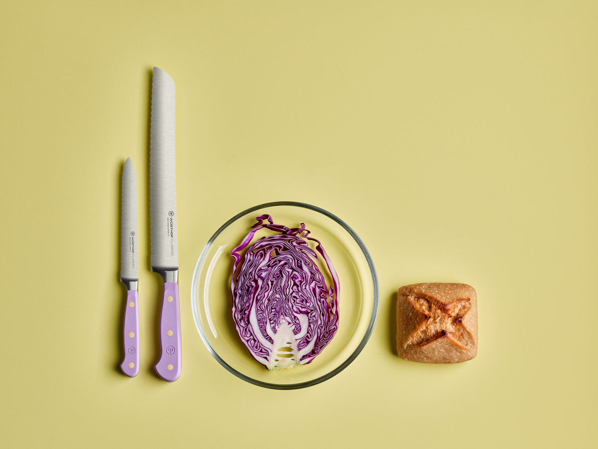 Classic 9" Precision Double-Serrated Bread Knife with a lavender handle displayed next to a smaller serrated paring knife, purple cabbage half, and bread roll.
