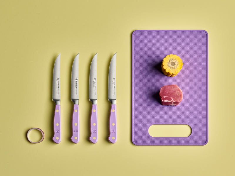 Classic 4-Piece Steak Knife Set showing blades with light purple handles featuring brass rivets, next to a purple cutting board with corn and meat.