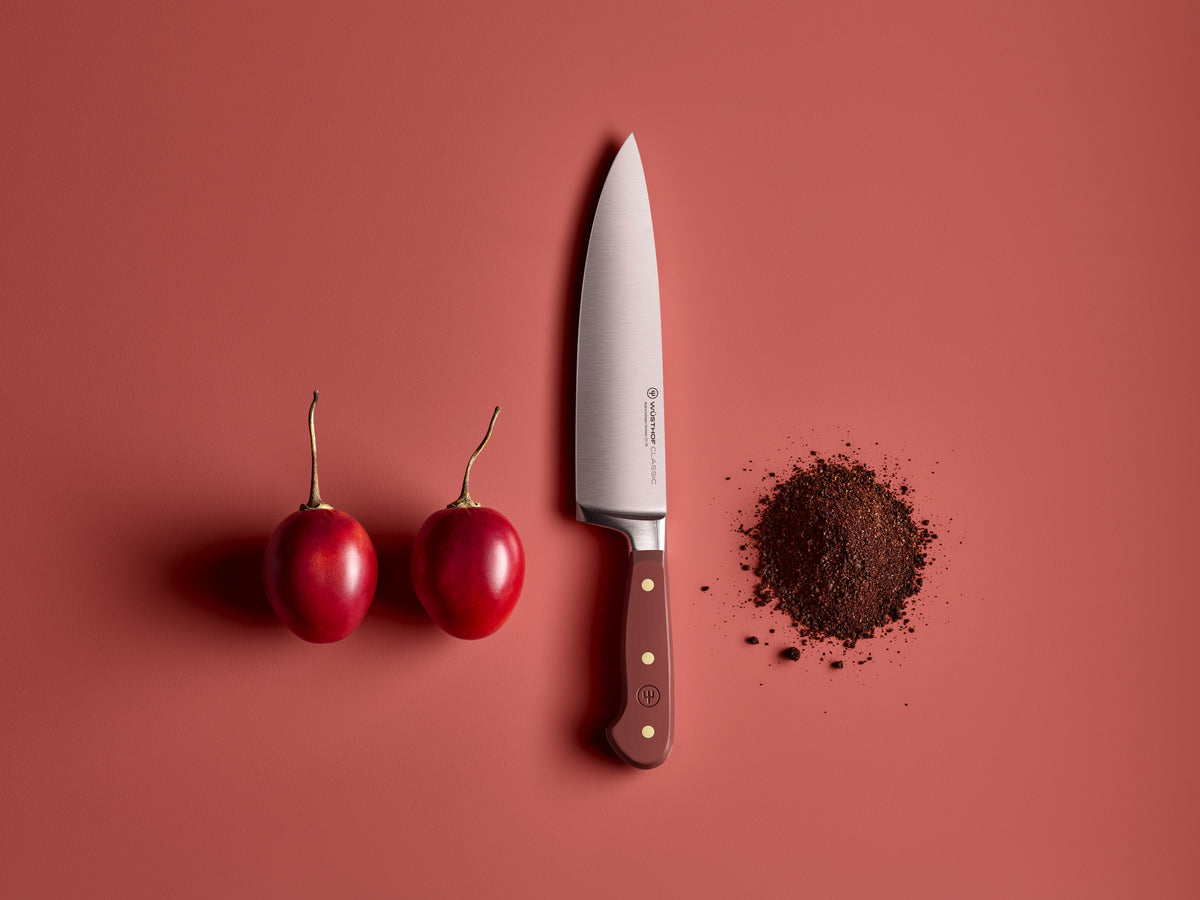 Classic 8" Chef's Knife with a polished forged German steel blade and smooth deep red handle, displayed beside two red cherry tomatoes and a pile of ground spices.