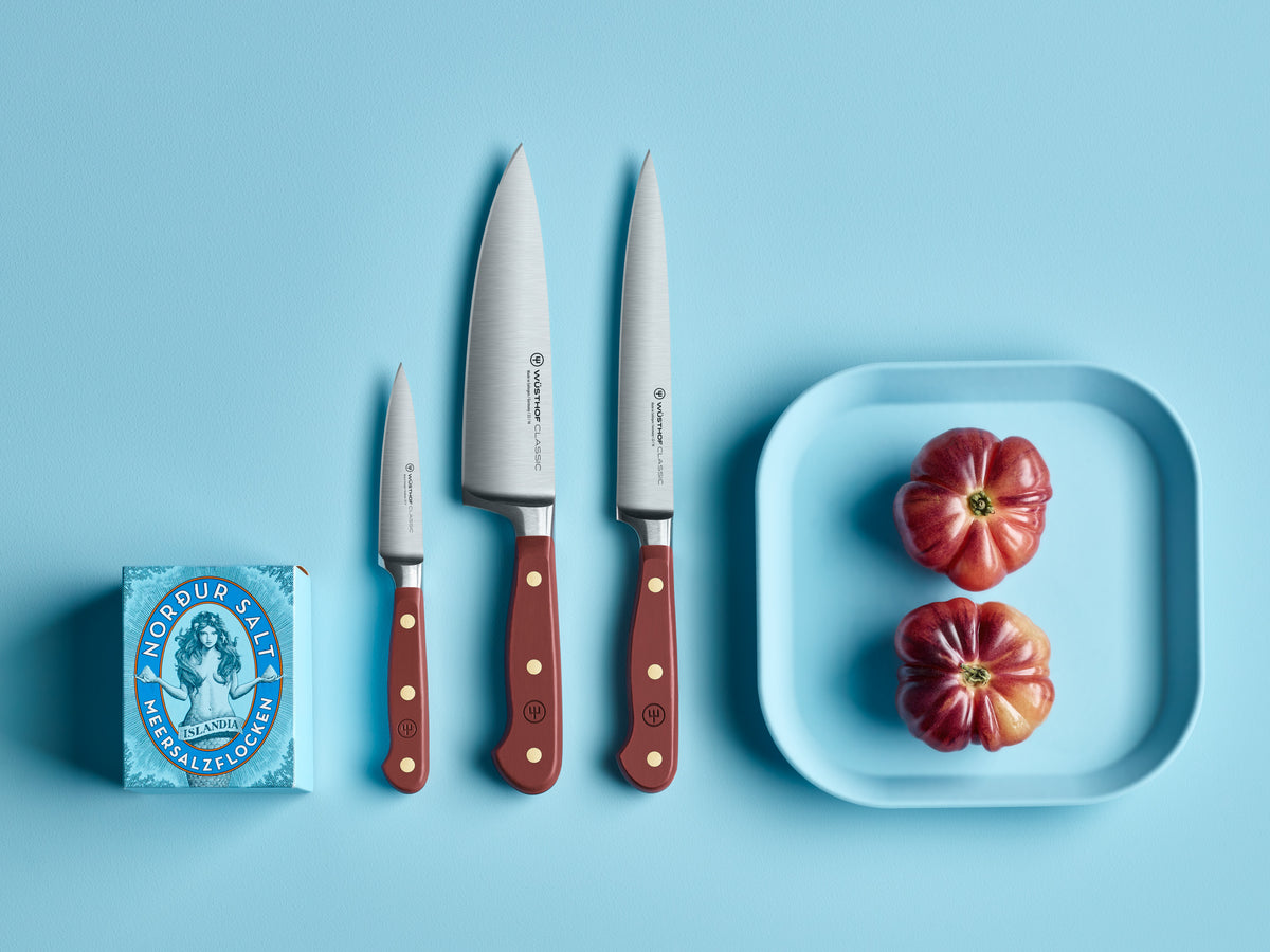 Three Classic 6" Utility Knives with burgundy handles and brass rivets arranged beside a blue plate with heirloom tomatoes and a salt box on a blue surface.