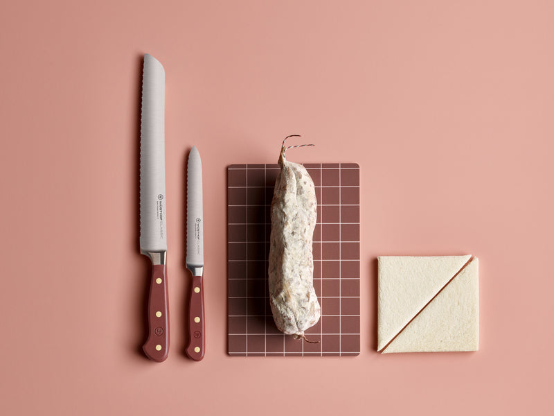 Classic 5" Serrated Utility Knife with a deep red handle and brass rivets shown alongside a smaller serrated knife, a cured sausage, and sliced bread on a pink background.