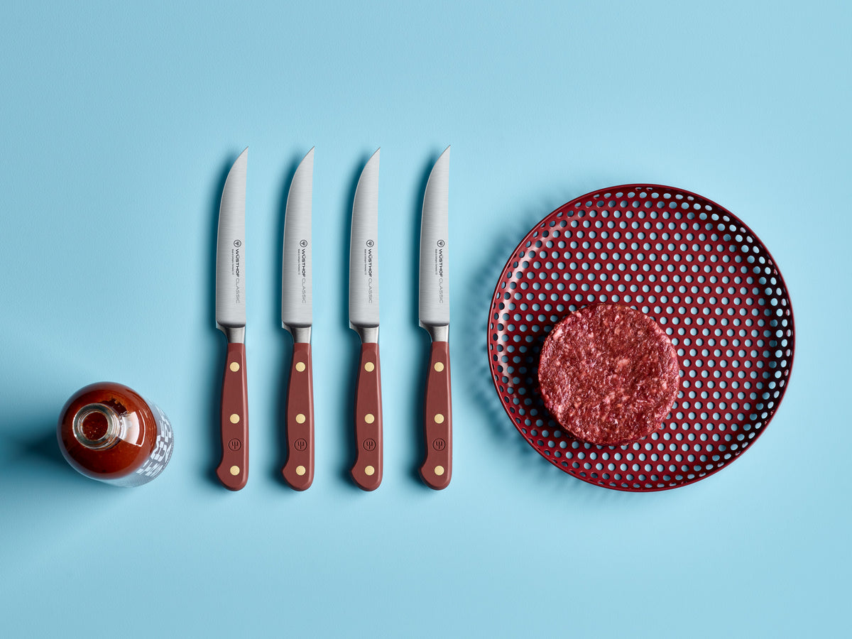 Classic 4-Piece Steak Knife Set with forged German steel blades and deep red handles featuring brass rivets, displayed next to a raw burger patty and sauce bottle.