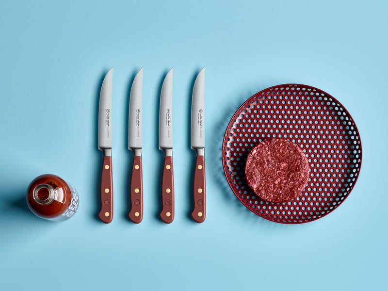 Four Classic 4 1/2" Steak Knives with polished red handles and brass rivets arranged beside a raw burger patty on a perforated red plate with a bottle of sauce nearby.