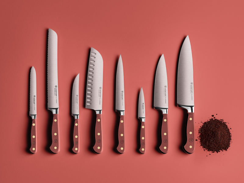 Classic 8" Chef's Knife displayed with a matched set of forged kitchen knives featuring polished German steel blades and deep red handles with brass rivets, arranged neatly on a matte red surface beside a small pile of ground spices.