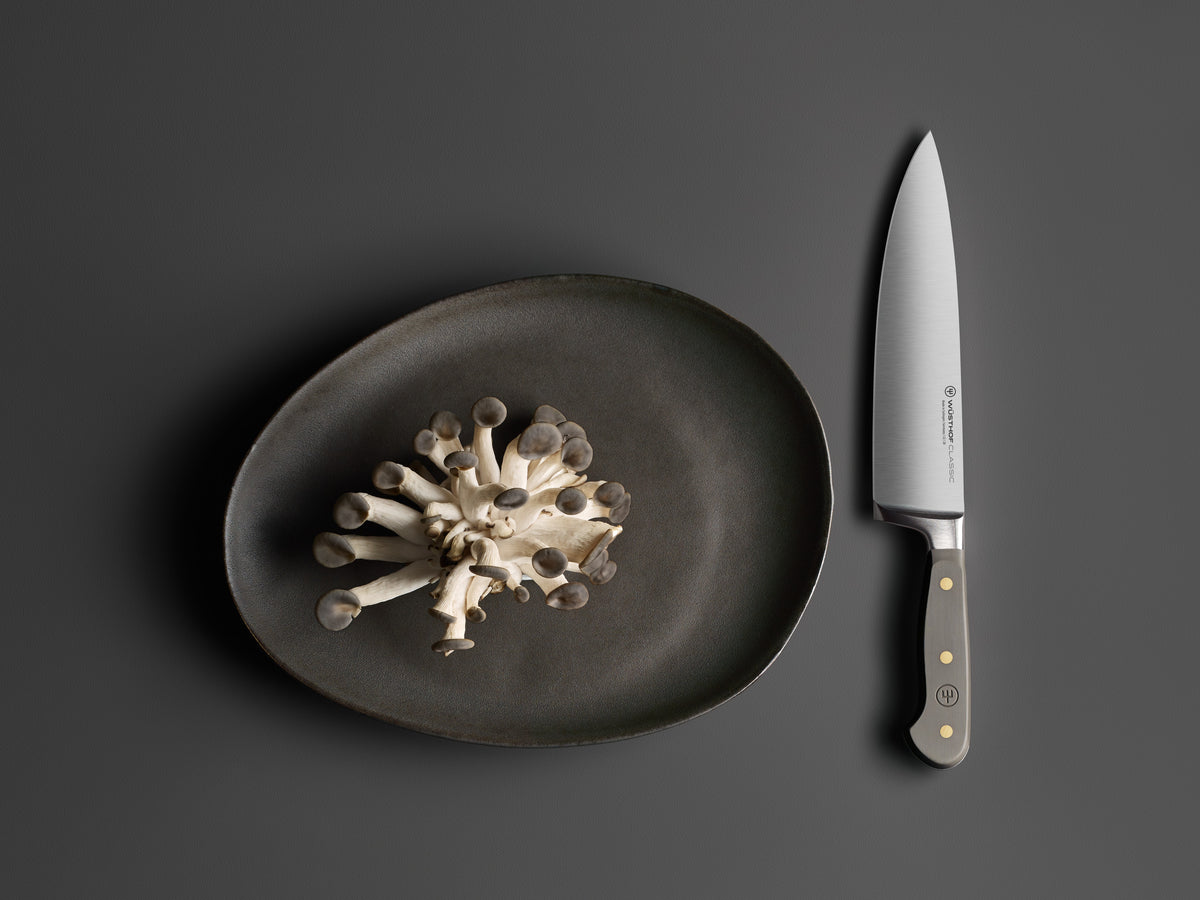 Classic 8" Chef's Knife with a polished forged German steel blade and matte dark handle featuring three brass rivets, displayed next to a cluster of fresh mushrooms on a dark plate.