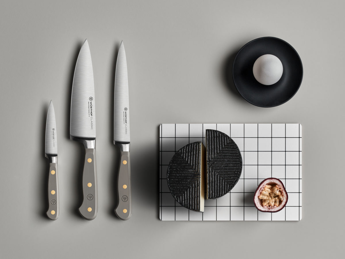 Three Classic 6" Utility Knives with gray ergonomic handles and brass rivets arranged beside black textured cheese and half a passion fruit on a white grid board.
