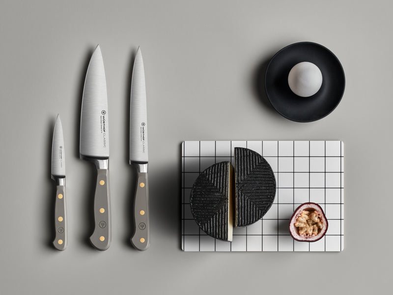 Three Classic 6" Utility Knives with gray ergonomic handles and brass rivets arranged beside black textured cheese and half a passion fruit on a white grid board.