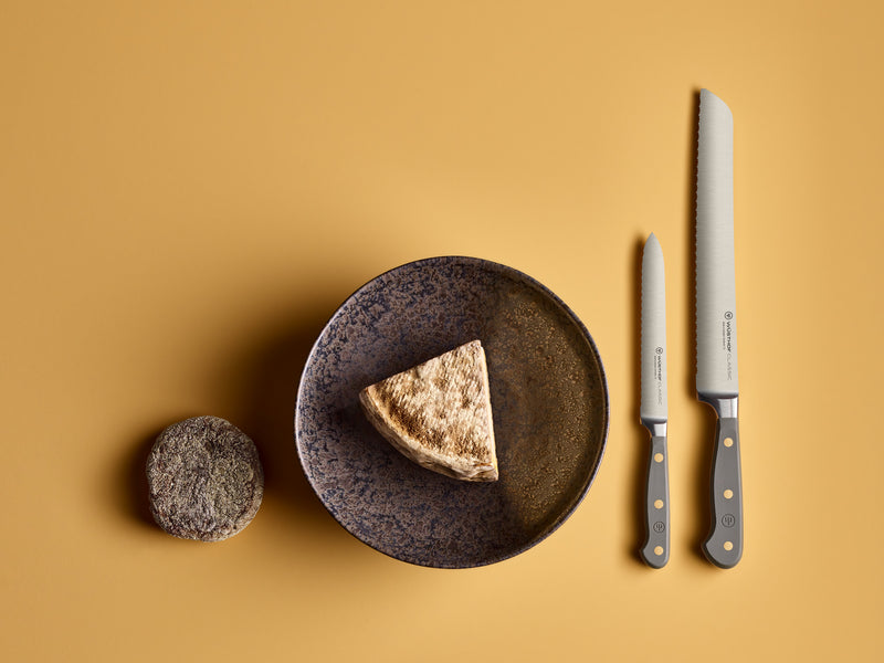Classic 9" Precision Double-Serrated Bread Knife with a taupe handle featuring brass rivets, displayed next to a matching paring knife and rustic bread on a brown ceramic plate.