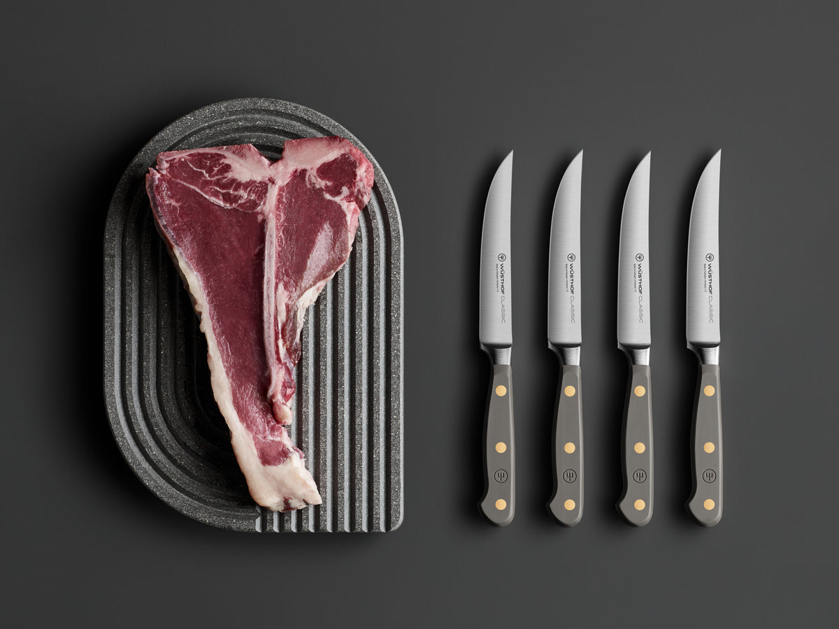 Classic 4-Piece Steak Knife Set featuring matte grey handles with brass rivets and sharp forged German steel blades, displayed beside a raw T-bone steak.