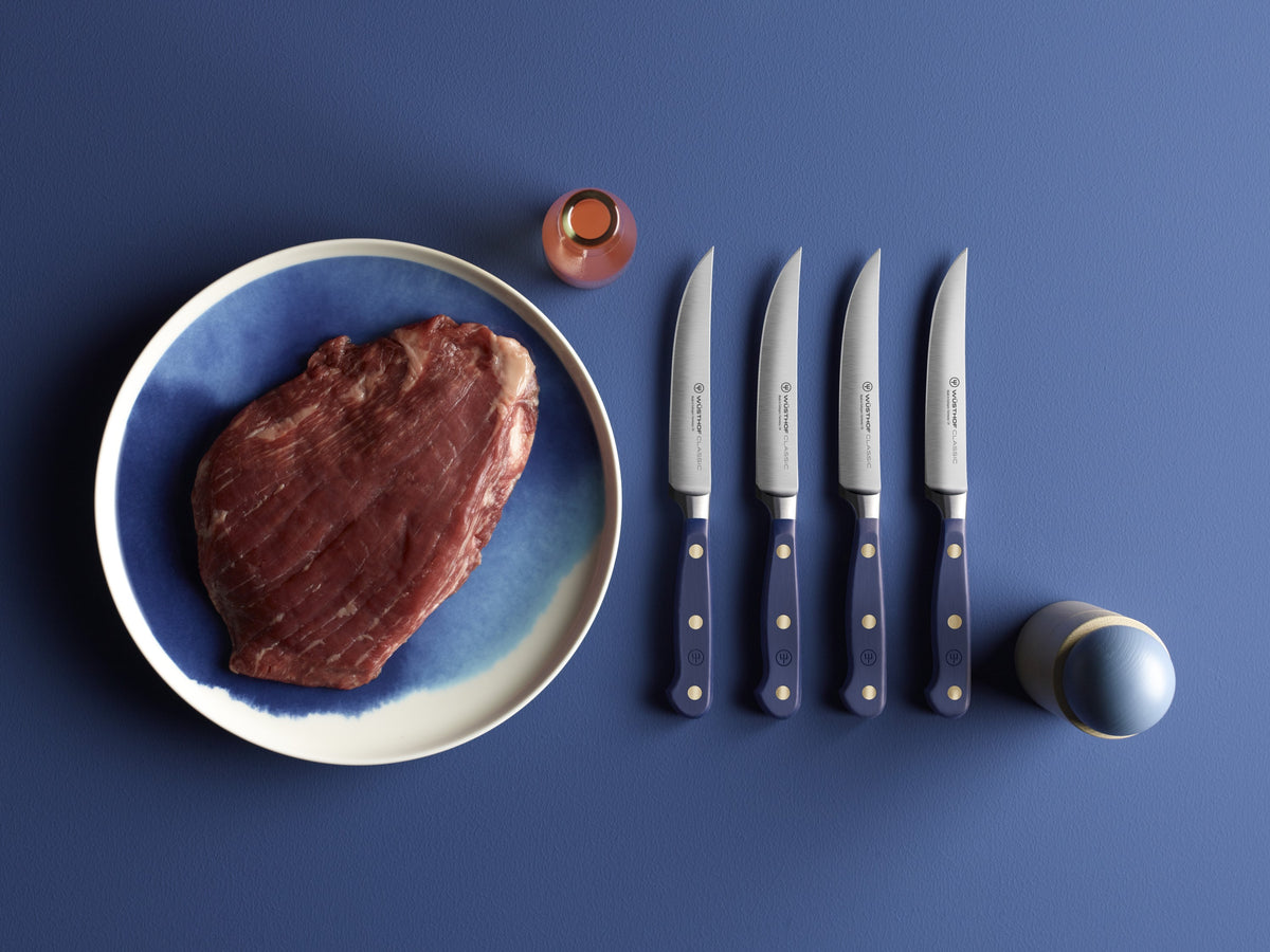 Four Classic 4 1/2" Steak Knives with navy blue handles featuring three brass rivets, arranged beside a raw steak on a blue ceramic plate.
