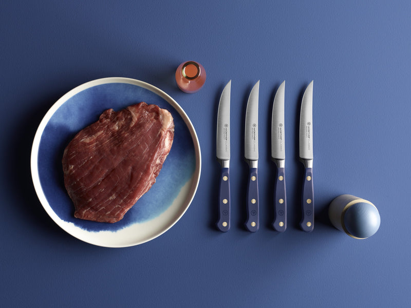 Four Classic 4 1/2" Steak Knives with navy blue handles featuring three brass rivets, arranged beside a raw steak on a blue ceramic plate.