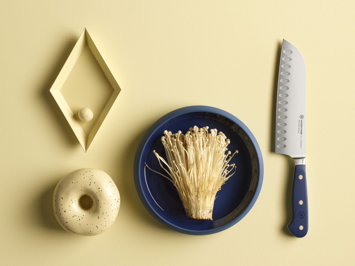 Classic 7" Hollow Edge Santoku with a navy ergonomic handle and hollow scalloped German steel blade next to enoki mushrooms on a blue plate.